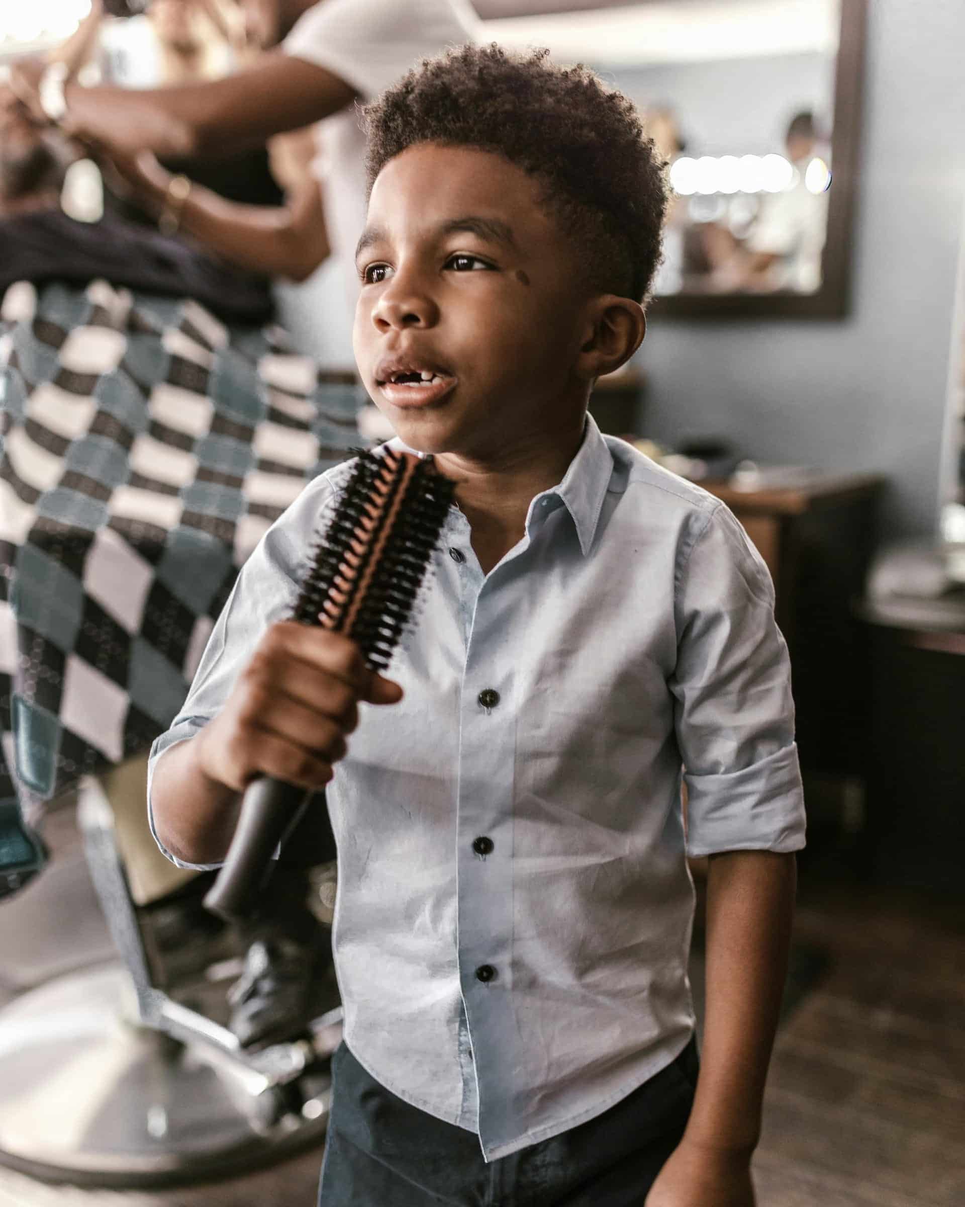 A young African American boy smiling and holding a hairbrush getting ready for Back to School with a curly top fade haircut from Floyds.